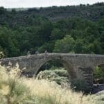 Three Slow Cyclists riding over a stone bridge over the Coa River in Portugal, with trees and hills in the background.